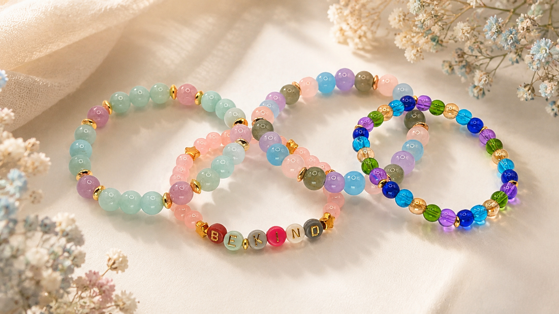 Four colorful beaded bracelets with gold accents and a "BE KIND" word bracelet arranged with delicate flowers on a softly lit background, showing how thoughtfully designed beaded jewelry can feel polished, expressive, and suitable for everyday wear.