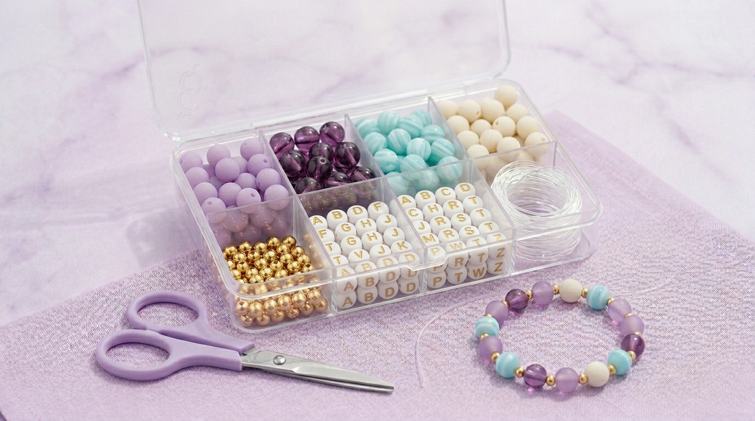 Organized bracelet making kit for beginners with glass beads, letter beads, golden spacers, and elastic cord arranged in a storage box, with a finished bracelet and scissors beside the kit on a marble table