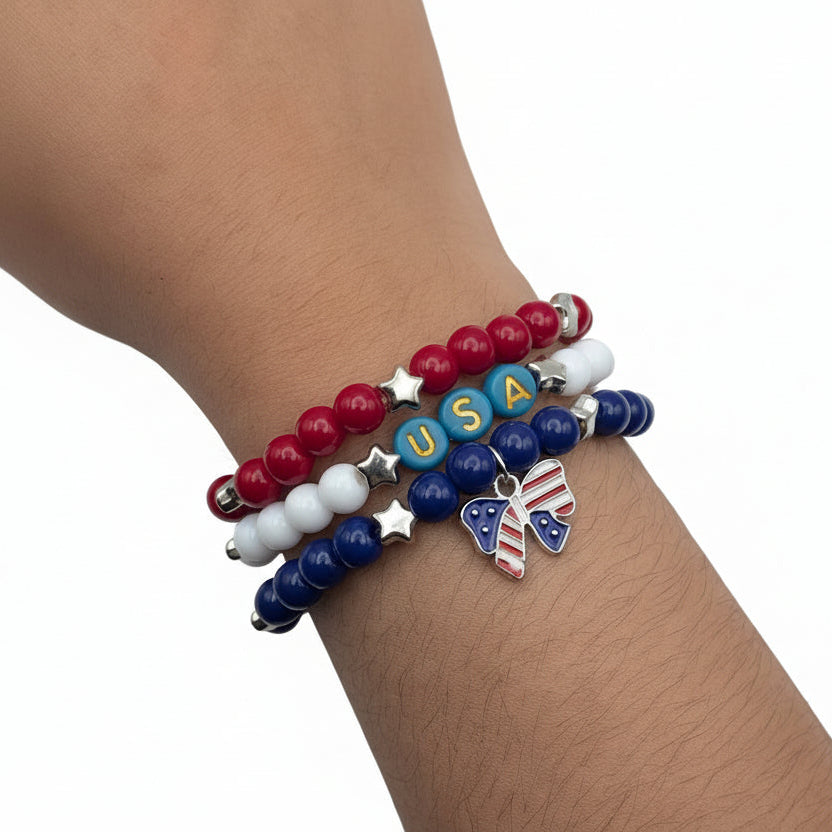 Stacked red, white, and blue patriotic bracelets worn on wrist featuring USA letter beads, star accents, and an American flag bow charm for Independence Day and summer celebrations